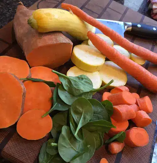A cutting board covered with fresh vegetables, including sliced sweet potato, carrot rounds and whole carrots, yellow squash slices, spinach leaves, and a kitchen knife resting nearby on a tiled surface.